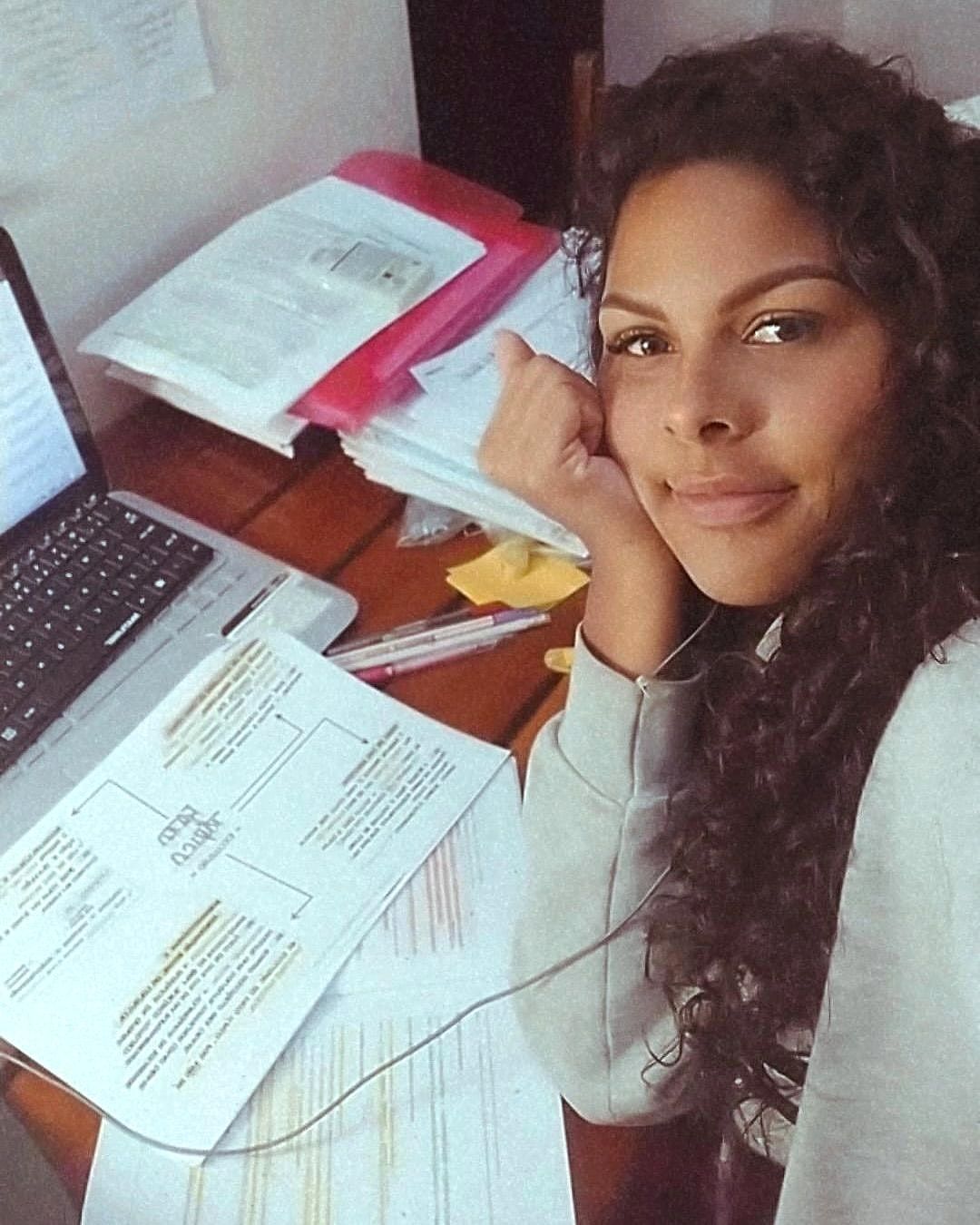 Ju sorrindo, apoiada em uma mesa cheia de papéis e anotações de estudo